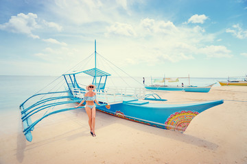 Enjoying travel and vacation in tropical paradise. Young pretty woman in hat and swimwear near the traditional boat on white sand beach.