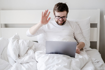Man communicates remotely through a video call from a laptop, he looks at the screen, smiles and waves hand while sitting in bed.