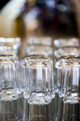 Rows of glasses in a bar waiting to be used. In the background there are bottles of alchocolic beverages.
