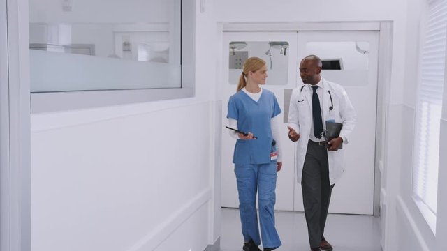 Doctor In White Coat And Nurse In Scrubs Having Discussion Over Digital Tablet In Hospital Corridor