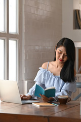 Beautiful woman reading a book while sitting in front a computer laptop that putting on the wooden table with comfortable living room as background.