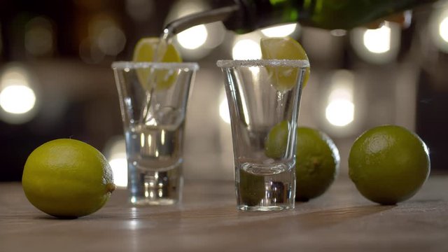 Close up bartender pouring tequila into glasses and sets fire to a strong alcoholic drink. Bar counter on the background of blurry light bulbs.