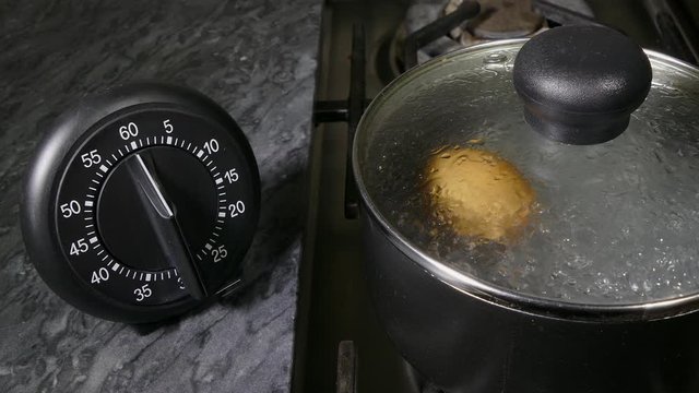 Close POV shot of a hen&rsquo;s egg being lowered into a pan of boiling water, followed by the lid to cover, then fingers setting the nearby manual mechanical timer for a few minutes to cook the egg.