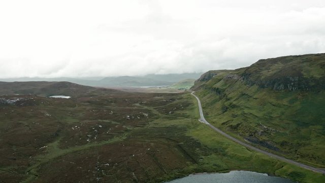 Aerial shot pans slowly around the rocky ridge of Knockan Crag in the Scottish Highlands. A road snakes off into. the distance. Peat bogs, grass and marshland are in the foreground.