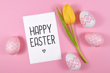 'Happy Easter' text card surrounded by easter eggs and single yellow tulip spring flower on pink colored background, flat lay