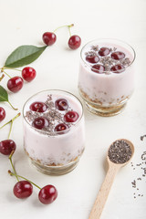 Yoghurt with cherries, chia seeds and granola in glass with wooden spoon on white wooden background. side  view.