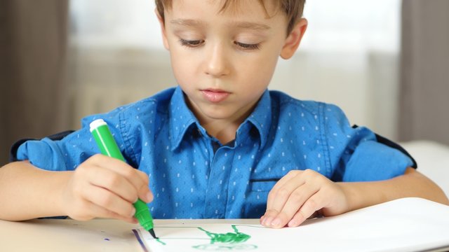 A little boy imagines, draws sitting at the table. The child draws bright felt pen