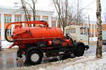 A truck with an orange tank with a white cab rides along a dirty city street in Russia in winter against the snow