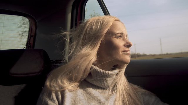 Young blonde woman smiles out car window as wind blows hair, slow motion