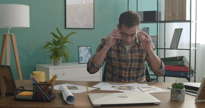 Young Caucasian man architect or designer taking on glasses and looking at the camera at the working table with drafts in the nice room.