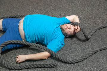 Overweight man is lying on the floor exhausted after performing battle rope exercise in the fitness...