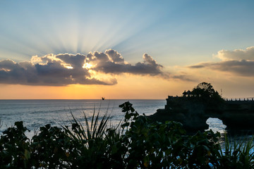 Cliffs in the nearby of Tanah Lot Temple, Bali, Indonesia. There is an arch in the water. The waves are splashing on the cliffs. Sun starts to set, hiding behind a cloud. Power of the nature