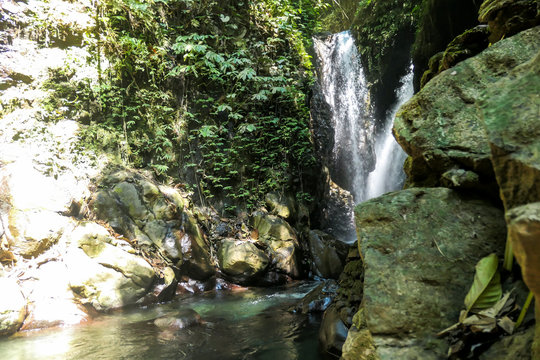 A view on the Gitgit Twin Waterfall in Bali, Indonesia. The waterfall is surrounded by rocks from each side. The slopes of the rocks are overgrown with plants. Wildness and inaccessibility of nature
