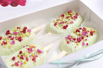 open box with four cake in the form of popsicle on a stick in light green glaze and flower and pistachio topping..