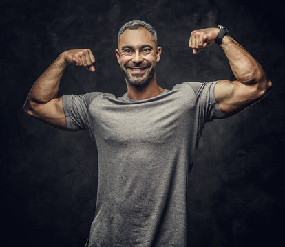 Strong, adult, fit muscular caucasian man posing for a photoshoot in a dark studio under the spotlight wearing sportswear, showing his muscles while smiling