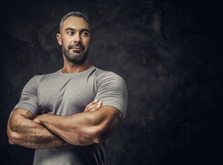 Strong, adult, fit muscular caucasian man posing for a photoshoot in a dark studio under the spotlight wearing grey sportswear, showing his muscles with arms crossed, looking confident and calm - Powered by Adobe