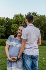 Young couple in love on a walk in the park, husband hugs pregnant wife by the belly, happy family