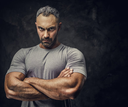 Serious, Adult, Fit Muscular Caucasian Man Coach Posing For A Photoshoot In A Dark Studio Under The Spotlight Wearing Grey Sportswear, Showing His Muscles With Arms Crossed Looking Angry