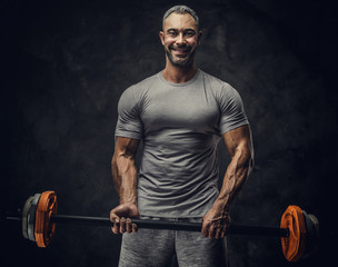 Naklejka na ściany i meble Strong, adult, fit muscular caucasian man coach posing for a photoshoot in a dark studio under the spotlight wearing grey sportswear, showing his muscles and holding a sport gear looking confident and