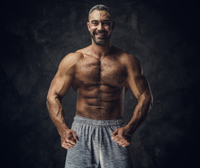 Fototapeta na wymiar Strong, adult, fit muscular caucasian man coach posing for a photoshoot without his shirt in a dark studio under the spotlight wearing sporty shorts, showing his muscles looking powerful and smiling