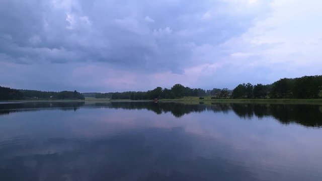 Gorgeous view of sunset on calm summer evening. Lake shore with green trees and plants reflecting in crystal clean mirror water surface. Sky covered with heavy thunder clouds.