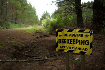 Be aware beekeeping activity ahead yellow and black sign warning sign beside an unpaved road track in Riverhead Forest, Kumeu, New Zealand.