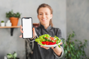 happy young woman shows a smartphone with a white screen on an outstretched hand and holds a plate with chopped vegetable salad in the other hand on the kitchen