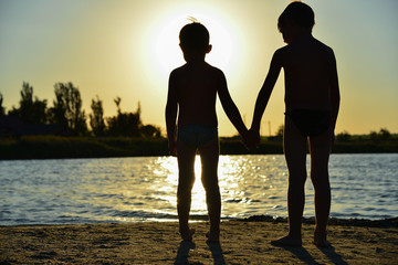 Two brothers are standing on the banks of the river and holding hands against the setting sun.