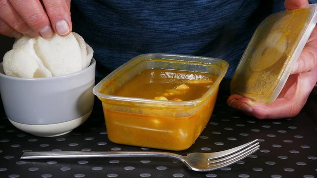 Cinemagraph – Close POV video of steam rising from a takeaway / takeout carton of Chinese king prawn curry, with a still image of a man’s hand holding the container lid and fingers on prawn crackers. 