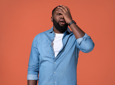 Tired man covers face with hand, stress. Photo of african man in casual outfit on coral background.