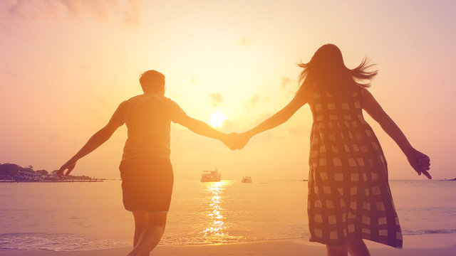 Happy couple holding each other hands walking to the sea at sunset.