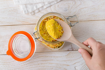 Woman hand taking universal seasoning with large wooden spoon from clip top glass jar on a white wooden kitchen table. Seasoning with salt and dry vegetables.