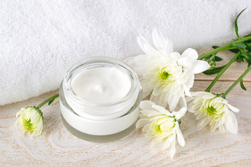 Fototapeta na wymiar Facial cream in an open glass jar next to fresh chrysanthemum buds and a white terry towel on a white wooden surface. Beauty, skincare and cosmetology concept.