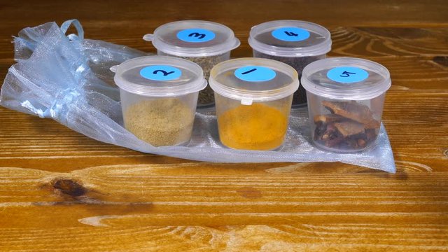 Close POV shot of a man&rsquo;s hands in a shop or market, setting out five small plastic pots on a pretty bag, containing samples of Indian spices and an identifying list.