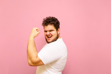 Joyful fat man showing biceps and looking into camera with smile on face isolated on pink...