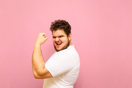 Joyful fat man showing biceps and looking into camera with smile on face isolated on pink background. Portrait of a big boy overweight, happy to win with his arm raised and posing for the camera.