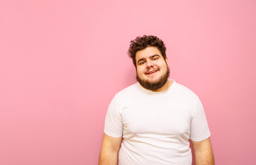 Portrait of a funny overweight guy on a pink background, wearing a white t-shirt and beard, looking...