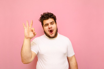 Happy young man with overweight and curly hair and beard shows OK gesture, looks into camera with joyful face. Positive totsun in white t-shirt shows OK gesture with his fingers. Isolated on pink. - obrazy, fototapety, plakaty