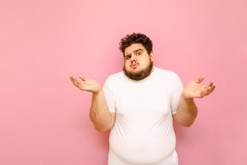 Portrait of a bewildered fat young man in a white T-shirt, looking into the camera and spreading his hands to the sides. Portrait of a puzzled overweight boy on a pink background. Copy space - obrazy, fototapety, plakaty