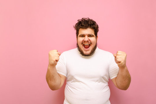 Portrait of a joyful guy in a white T-shirt and overweight rejoices the victory of his beloved team on a pink background. Happy fat cheerful man with beard raised his hands up for joy. Copy space