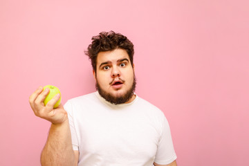 Surprised young overweight man stands on pink background with green apple in hand, looks into...