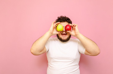 Happy fat guy in white t-shirt stands on pink background and holds 2 apples at eye level and...