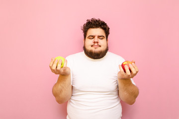 A funny fat young man in a white T-shirt stands with two apples in his hands on a pink background...