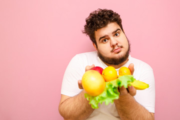 Closeup portrait of a funny young man with beard and curly hair holding fruits and vegetables in his hand and looking into the camera. Fat young guy holding healthy food in his hand, diet concept.