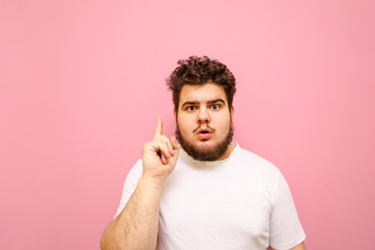 Serious young overweight man in white t-shirt, isolated on a pink background, looks into the camera with a surprised face and shows finger up. Funny fat guy has an idea, stands on a pink background