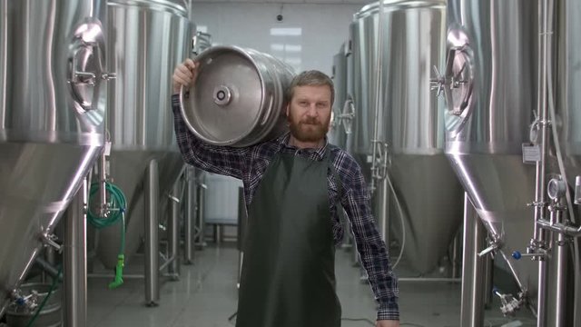 Worker Male brewer carries a keg filled with beer passing beer tanks