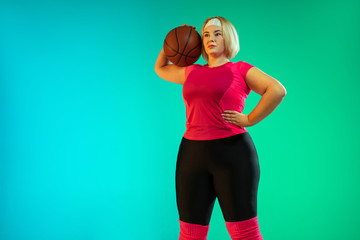 Young caucasian plus size female model's training on gradient green background in neon light. Doing workout exercises with ball, basketball. Concept of sport, healthy lifestyle, body positive