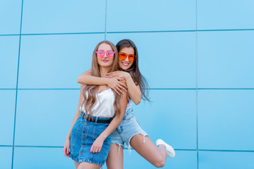 two young hipster woman friends in retro neon sunglasses standing and smiling over blue wall