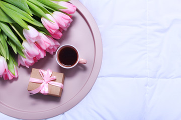Tray with cup of coffee,gift box and bouquet of pink tulips on white sheet on bed.Flat lay,spring, concept woman's or mother's day top view
