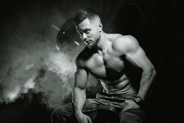 Fototapeta na wymiar Bodybuilder posing on black background. Sitting on bench after training. Muscular man in gym. Black and white photo